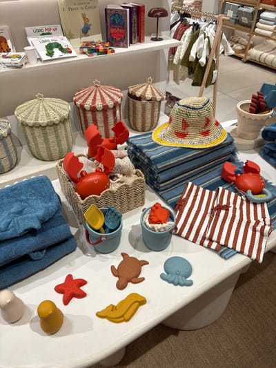 The image depicts a retail store display in Leganés, Spain, featuring children's items. In the foreground, a white, rounded display table occupies the majority of the frame. On the table's left side, a stack of dark blue folded towels, one possibly with an embroidered whale tail or shark fin, sits next to three cylindrical woven baskets with pointed, star-adorned lids. These baskets are striped in green/white, red/white, and light brown/white, respectively.

In the center of the table, a rectangular woven basket contains two large red plastic crab claws and a mesh bag holding additional red crab-shaped toys. Adjacent to this, a stack of blue and white striped fabric with frayed edges is topped by a straw sun hat featuring red boat patterns, blue bands, and a yellow brim. To the right of the hat, two small light blue silicone buckets hold yellow shovels and blue rakes; one bucket also contains a red crab-shaped toy. Beside these, a folded red and white striped children's garment, possibly shorts or swim trunks with red ties, is displayed next to a red crab-shaped teether or rattle. Further forward on the table, an assortment of silicone bath or beach toys includes a red starfish, a brown turtle, a blue octopus, a yellow seal-like figure, a yellow bird, and a beige rounded figure.

In the mid-ground, behind the display table, a clothing rack holds various children's garments in light, muted colors. Above this, on a white wall shelf, an array of children's books is visible. Titles include "El Cuento de Perico el Conejo" (The Tale of Peter Rabbit), a "Harry Potter" book, and several "LIBRO POP-UP" (Pop-Up Book) editions, one of which features "The Very Hungry Caterpillar." A small, dark red mushroom-shaped table lamp and small, colorful blocks are also on the shelf. The scene is well-lit, suggesting an indoor retail environment. No people are visible.