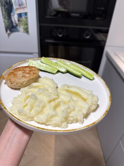 In the city of Postol, Russia, a white plate with a brown rim holds a meal. The plate is positioned on a light-colored countertop. On the plate, there is a golden-brown patty on the left, and a serving of fluffy mashed potatoes to the right and front. Three sliced cucumbers are arranged in a row behind the patty. The background is blurred, showing a modern kitchen environment with kitchen appliances. The image is taken from a slightly elevated angle, with a hand holding the plate visible in the lower left corner.