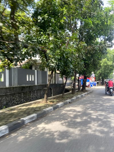 The image shows a street in Kecamatan Cimahi Selatan, Indonesia, located at latitude -6.909 and longitude 107.568. The street is lined with trees on the right side and a gray wall with white stripes on the left side. There is a concrete curb with a white and black painted stripe separating the road from the sidewalk. A person is riding a motorcycle on the road toward the right side of the image. The motorcycle is red with a black seat. The person is wearing a red shirt. The background is blurred.