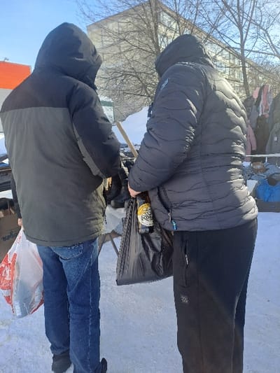 The scene depicts an outdoor winter environment in Bolshaya Elhovka, Russia. Two individuals are observed from behind, standing on snow-covered ground under a clear sky. In the background, a multi-story building with numerous windows is visible, along with bare tree branches. An orange structure is partially visible on the far left.
The individual on the left wears a two-tone hooded jacket (dark upper, gray lower), blue denim trousers, and dark footwear. They hold a transparent plastic bag with red markings in their left hand, which appears to contain items wrapped in plastic.
The individual on the right wears a dark, hooded, quilted jacket and black athletic pants with a brand logo on the left thigh. They hold a black plastic bag in their left hand, from which a brown glass bottle with a yellow label, identifiable as "Kozel" beer, protrudes.
Both subjects are positioned facing a partially visible outdoor vendor's display or market stall, which holds various objects. Between them, a large white, rectangular object with a wooden handle is visible. Further to the right in the background, clothing items are hanging on a rack.