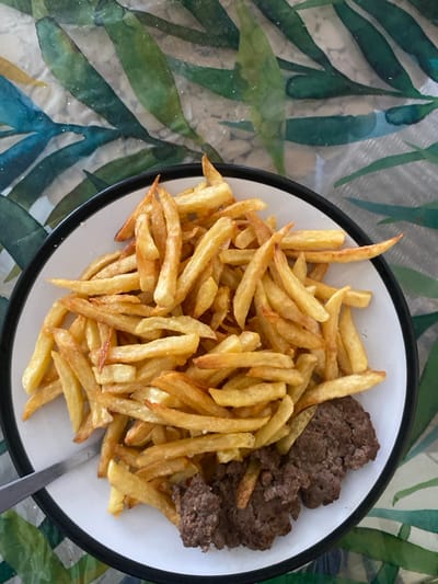 The image displays a meal served on a white plate with a black rim. The plate contains a substantial portion of golden-brown french fries occupying most of its area. To the bottom right, three irregular clumps of cooked ground meat, brown in color, are visible, partially obscured by the fries. A silver-colored fork handle is positioned at the bottom left, extending beneath the fries on the plate. The plate rests on a surface covered with a patterned tablecloth or placemat featuring large green and teal leaf motifs against a lighter, textured background. The scene is located in Capesterre-Belle-Eau, France. No individuals or active interactions are depicted.