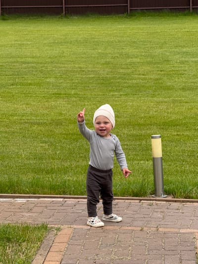 In this image, a baby is standing on a paved pathway next to a lush green lawn. The baby is wearing a white beanie, a striped long-sleeved shirt, dark pants, and white sneakers with colorful accents. They are smiling and pointing with their left index finger. A metal pole with a yellow and gray cylindrical top is visible in the background, partially obscured by grass. The setting appears to be outdoors, possibly a backyard or park, given the lawn and the paved path. The lighting suggests it's daytime, and the clear visibility indicates fair weather. The image seems to capture a candid moment of the child exploring their surroundings. The location context provided is Борисьево Днп, Russia. There is no visible text in the image.