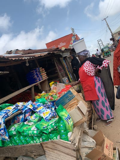 The image depicts a bustling outdoor market scene in Jos, Nigeria, focusing on a woman carrying a white plastic tub on her head. She is dressed in a red apron over a white and red patterned top and a long, patterned pink and blue skirt. Beside her, a stall displays an abundance of colorful detergent packets, primarily green and blue, along with a pile of red chilies and bars of soap. In the background, other market-goers, a green tricycle, and overhead power lines suggest a lively urban environment. The sky above is a mix of blue and scattered clouds, indicating daylight.
