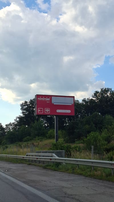 A red billboard advertising "TOYOTOMI air conditioners Japan" is mounted on a tall metal pole beside a highway. The billboard displays the brand name, "air conditioners Japan," an "A+++" energy efficiency rating, a Wi-Fi symbol, and a search icon with the website "www.bgr.bg." A white air conditioning unit is depicted on the right side of the billboard. The billboard is positioned against a backdrop of lush green trees and a cloudy sky. The foreground shows a section of the highway with a metal guardrail running along its edge. The image appears to have been taken in the city of Mirovo, Bulgaria.