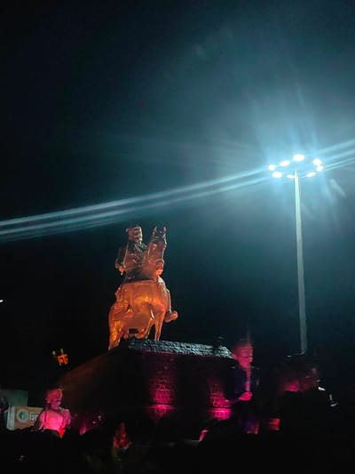 The image shows a night scene in Birsingpur, India, featuring a prominent equestrian statue illuminated by bright spotlights. The statue depicts a historical figure on horseback, rendered in a golden hue, and is positioned atop a dark, textured pedestal. Below the pedestal, pink lights cast a glow on what appears to be a crowd gathered at the base, with several individuals visible. A tall lamppost with multiple bright lights is visible on the right side of the frame, casting strong beams that create lens flare and illuminate the sky. The overall atmosphere is dark, with the bright lights creating dramatic contrasts and highlighting the central monument and the surrounding activity.