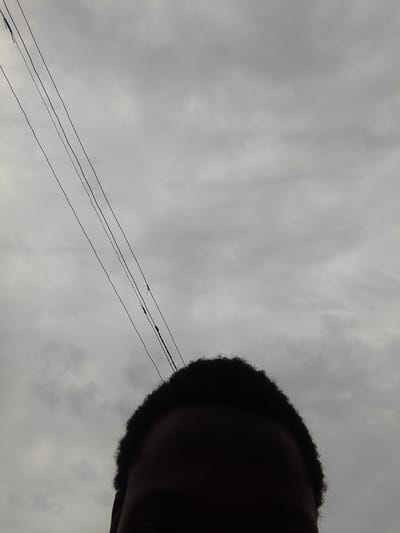 The image shows a close-up view of a person's head and hair against a cloudy sky. Three power lines are visible, running diagonally from the upper left to the middle right of the frame. The person's face is mostly obscured in shadow, with only the outline of their forehead and curly hair visible. The background consists of overcast, grey clouds. The setting is the city of Alagbede, Nigeria.