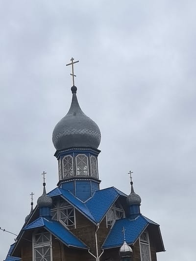 The image shows the top section of a wooden Orthodox church with bright blue roofs and gray domes. Multiple crosses top the domes. The main dome is large and bulbous, with a smaller, onion-shaped dome below it. The wooden walls are visible between the windows and roofs. The sky is overcast and gray. The church appears to be located in a rural area, consistent with the Камянюцкі сельскі Савет, Belarus. There are no people visible in the image. The overall impression is one of religious architecture against a somber sky.