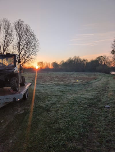 The image depicts a sunrise scene near Zetiovo, Bulgaria, showing a field covered in frost. The sun is rising on the horizon, partially obscured by trees. A utility terrain vehicle (UTV) is parked in the foreground. The sky is a gradient of colors from pale orange to light blue. The field has a yellow golf ball on the frosted grass.