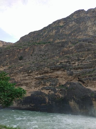 This media file depicts a dramatic natural landscape, likely a canyon or gorge, with a powerful river flowing at its base. The dominant subject is a massive, rugged mountain or cliff face that rises steeply from the water's edge. The rock formation is layered and textured, showing signs of erosion and weathering, with various shades of brown, tan, and dark grey. Sparse vegetation, including green shrubs and a few trees, clings to ledges and the lower slopes of the mountain.
The river is a significant element, appearing turbulent and fast-moving, with white foam and ripples indicating strong currents, possibly a rapid or waterfall just out of frame to the right. The water is a greenish hue. On the left side of the frame, the lush green foliage of a tree or bush frames the lower portion of the scene, providing a contrast to the rocky terrain.
The sky above is overcast with diffused light, suggesting it might be a cloudy day or early morning/late afternoon. There are no people or man-made objects visible in the image, emphasizing the wild and untouched nature of the setting. The overall mood is one of awe and the raw power of nature. The location context provided places this scene in Untsukul', Russia, suggesting a mountainous or riverine region within that area. There is no visible text within the image.
