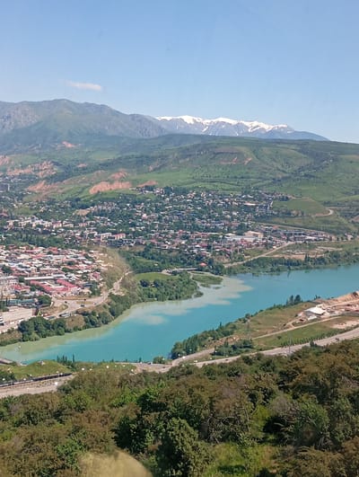 The image captures a high-angle view of Chinarkent, Uzbekistan. A town with buildings is spread across a valley. A river runs through the scene, its color varying from turquoise to a muddy brown. Mountains with snow-capped peaks are visible in the background. The foreground features trees. A road runs beside the river.