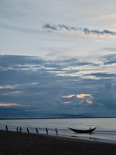 The image depicts a beachfront scene at dusk in Uleekareung, Indonesia. In the foreground, a sandy beach is visible, with a few scattered pieces of litter. A group of seven fishermen, silhouetted against the water, are engaged in pulling a fishing net from the sea. They are spaced out along the water's edge, each straining to pull the heavy net. Further out in the calm water, a lone wooden boat is partially submerged, with a person standing near it. The background is dominated by a vast expanse of ocean, with a distant shoreline featuring rolling hills and mountains. The sky is filled with dramatic clouds, tinged with the soft colors of sunset, creating a moody and atmospheric backdrop. The overall impression is one of hard work and the vastness of nature.