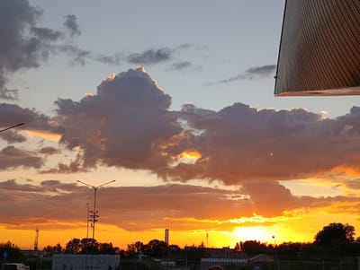 This media file depicts a dramatic sunset or sunrise over an urban or industrial fringe landscape in Bolshoye Savino, Russia. The sky is the dominant subject, filled with a dynamic display of colors and clouds. The upper portion of the sky is a pale blue, gradually transitioning to vibrant oranges and yellows near the horizon, suggesting the golden hour.
Numerous clouds are scattered across the sky; some are large and dark grey, while others have bright, glowing edges where they are illuminated by the unseen sun. A particularly prominent, towering cloud mass is located in the upper-mid section, also featuring a radiant, sun-kissed top.
Silhouetted against the brilliant sky are various features of the landscape below. Dark, leafy trees line the horizon, interspersed with industrial-looking structures such as tall towers or chimneys. Two distinct streetlights or lampposts stand in the mid-ground, their intricate forms starkly outlined by the intense light behind them. Hints of buildings or low structures are visible at the very bottom.
A notable detail is a large, dark, angled structure in the upper right corner, with a textured, vertically striped surface. It appears to be an architectural element, possibly an overhang or part of a building, extending into the frame and partially obscuring the sky. No discernible text is visible. The overall mood is one of natural beauty and urban melancholy, characteristic of an evening sky.