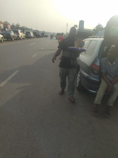 The scene takes place on a paved road in Maraba, Nigeria (latitude: 9.024, longitude: 7.573).  A man in a black shirt and khaki pants walks toward the right of the frame, carrying a tray of snacks. To the right of the man, a silver car is parked on the side of the road, and another man is seated on its trunk.  The man on the trunk wears khaki pants and a light-colored shirt. Both men are wearing sandals. The sun is shining brightly in the background.