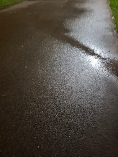 The image depicts a close-up, ground-level view of a wet asphalt surface, likely a path or road, with some grass visible on the right side and in the upper left corner. The asphalt is dark and reflective, showing numerous small, bright white highlights that indicate the presence of water, likely from rain or dew. There are also some subtle shadows or lighter patches on the surface, suggesting variations in moisture or light. The overall impression is one of an outdoor urban environment after or during precipitation, with no people or specific objects clearly identifiable. The location is specified as Novlyanka, Russia, which suggests a temperate climate where such weather conditions are common.