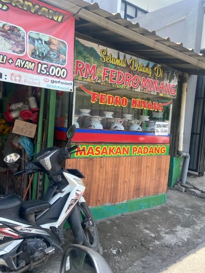 The image shows a storefront in Kecamatan Dayeuhkolot, Indonesia, with a sign that reads "Selamat Datang Di RM. FEDRO MINANG" in red letters on a yellow background. Below the sign is another, smaller sign that reads "FEDRO MINANG" in black letters on a white background. Below this, the window displays various bowls and plates, and the sign reads "MASAKAN PADANG" in green and yellow letters on a red background. A white motorcycle is parked in front of the store. The storefront has wood paneling and is partially obscured by a green wall. The scene is taken from the perspective of a person standing near the motorcycle, looking towards the storefront. The image captures a moment in the daily life of a small business in Indonesia.