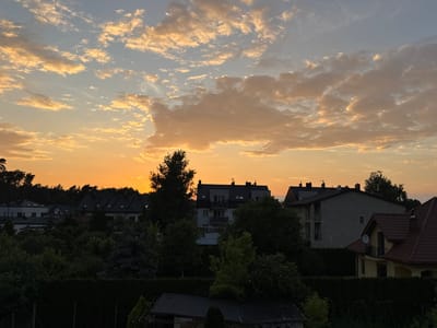 The image depicts a suburban residential area in Marki, Poland, during a sunset. The sky is filled with clouds illuminated by the setting sun, casting a warm, orange and yellow glow. In the foreground, dense green foliage and hedges form a barrier, obscuring the lower portions of buildings and gardens.
Behind the foliage, several residential buildings are visible. These include multi-story apartment buildings with dark tiled roofs and visible windows, as well as smaller houses with red tiled roofs and balconies. Some buildings have satellite dishes mounted on their exteriors. The architecture suggests a mix of modern and older styles common in European suburbs.
The overall impression is of a peaceful evening scene in a developed, yet green, urban environment. There are no people or animals visible in the frame. The lighting indicates it is either early morning or late evening, with the sun low on the horizon. The vegetation appears well-maintained, suggesting a residential area.