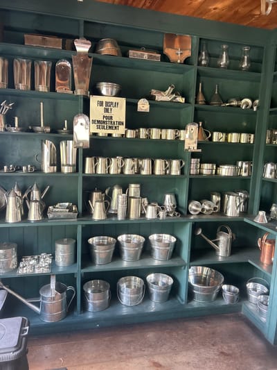 This is a well-lit, eye-level photograph of a display of metal kitchenware and garden tools on dark green shelves. The display is likely within a shop or museum in South Dundas, Canada, given its traditional, rustic aesthetic and the collection of vintage items.

On the top shelf, several silver-colored pitchers and beakers are arranged. To the right, a small, rectangular metal box with a hinged lid is visible, alongside a domed metal lid. Further to the right, several clear glass hurricane lamps are displayed.

The second shelf from the top features a collection of conical metal funnels, some stacked. Next to these, a series of silver-colored cups and mugs are neatly lined up. A sign with French and English text, "FOR DISPLAY ONLY / POUR DEMONSTRATION SEULEMENT," is prominently displayed in the center of this shelf.

The third shelf holds a variety of metal pitchers, kettles, and pots. Some are with spouts and handles, suggesting they are for pouring liquids. Several stacking metal containers are also present.

The fourth shelf is filled with more metal pitchers and kettles, some larger than those on the shelf above. A set of rectangular metal trays are stacked in the center.

The bottom shelf displays various sizes of galvanized metal buckets, some with handles. A metal watering can with a long spout is positioned on the left. To the right, more buckets are arranged, and a copper-colored pot is visible on the far right.

The shelves are made of dark green painted wood, and the overall impression is one of an organized and curated collection of antique or replica metal housewares and gardening implements. The background shows a wooden ceiling, suggesting the interior of a building with rustic architecture. There are no people visible in the image.