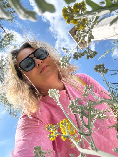 The image, taken in Bol'shoi Utrish, Russia, presents a low-angle outdoor perspective looking up at a blonde woman and surrounding vegetation against a blue sky.

The woman, with wavy, blonde hair appearing somewhat wind-blown, occupies the central and lower-mid portion of the frame. She wears black, rectangular-framed sunglasses with a light pink beaded chain, adorned with small gold accents, attached to the temples. A small stud piercing is visible in her left nostril. Her lips are closed, and she looks directly downwards towards the camera with a subtle expression. She is dressed in a long-sleeved, pink top with a faint, lighter pink pattern. A yellow strap, possibly from a bag or garment worn underneath, is visible over her right shoulder.

The foreground and sides of the image are filled with various plant life. Prominently featured are silvery-green plants with fuzzy, lobed leaves and clusters of small, round, pale green buds, some beginning to reveal tiny yellow florets. Additionally, clusters of bright yellow composite flowers on thin green stems are visible, particularly in the upper right and lower center. In the upper left quadrant, fine, needle-like green foliage, indicative of a coniferous tree or shrub, is partially visible.

The background consists of a bright blue sky with scattered white cumulus clouds. In the upper right corner, a white or light-colored wooden structure, appearing to be an exterior corner or roofline of a building, is sharply angled against the sky.