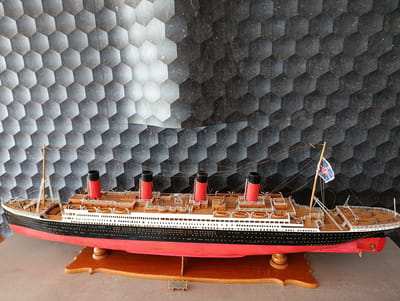A model of the RMS Titanic, painted red, black, and white with four funnels, is displayed on a wooden stand. The ship model is positioned on a gray surface in front of a black and gray hexagonal wall. The model features a flag with a Union Jack design. A small plaque on the stand reads "TITANIC 1911 G705 02 91". This scene is located in the city of Glifada, Greece.