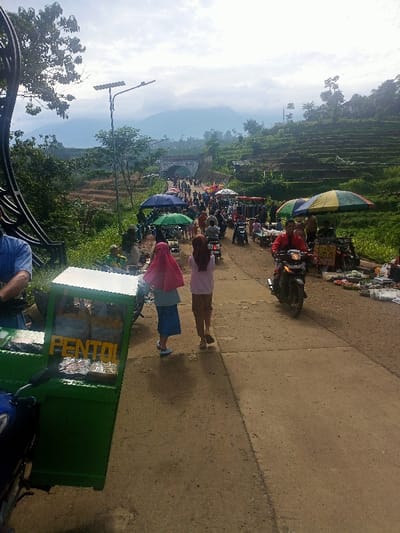 The image depicts a lively open-air market scene along a paved road in a hilly, mountainous region of Kecamatan Pangalengan, Indonesia.
In the immediate foreground on the left, a vibrant green food cart is visible, with the word "PENTOL" clearly printed on its side, indicating it sells an Indonesian street food. Partially visible next to the cart is a person in a blue shirt. In the center of the road, two women walk away from the viewer; one wears a pink hijab and a blue skirt, while the other is dressed in a light top and brown trousers.
The road is bustling with activity, featuring numerous people walking and several motorbikes navigating through the crowd. Market stalls with colorful umbrellas (blue, green, yellow, white, and red) line both sides of the road, displaying goods for sale. A man in a red jacket rides a motorbike towards the viewer on the right side of the road. Further down the road, a significant gathering of people is visible near what appears to be an archway or bridge structure. Solar-powered streetlights are also present along the path.
The background showcases lush green terraced hillsides, characteristic of agricultural landscapes, leading up to large mountains partially covered by clouds. The sky is bright but mostly overcast, indicating a daytime setting. The scene conveys a busy, vibrant local market atmosphere set against a scenic, rural backdrop.