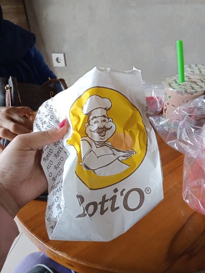A close-up indoor shot captures a person's hand holding a white paper bag from "Roti'O," a popular bakery chain. The hand, adorned with red nail polish, firmly grips the bag, which features a cheerful cartoon chef character holding a bun on a yellow circular background. The brand name "Roti'O" is prominently displayed in brown letters below the logo, and is also visible in a repeating pattern on the side of the bag. Inside the bag, a golden-brown baked good is partially visible through the chef's arms.
In the background, out of focus, another person wearing a blue hijab is partially visible, along with a dark-colored shoulder bag. On the right side of the wooden table, several plastic cups with green straws and sealed lids, characteristic of bubble tea or similar beverages, are bundled in clear plastic bags, suggesting multiple purchases. A white electrical outlet is visible on the light-colored wall in the background. The scene suggests a casual outing or meal in a relaxed setting, possibly a cafe or food court in Kecamatan Panji, Indonesia. The focus is on the Roti'O bun, implying it is the subject of immediate interest.