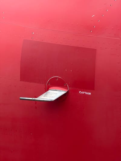 The image displays a close-up, vertical view of a vibrant red metallic surface, consistent with an aircraft fuselage, located in Tremblay-en-France, France. A silver-colored, metallic retractable step is deployed horizontally, extending towards the left from a circular base secured by four visible screws. The step's upper surface is stamped with instructions including "STEP", "PULL OUT TO USE", "PUSH IN TO STOW", and "MAX. WEIGHT 120 KG", along with identifying marks like "P/N 532320-101" and "CAGE 35738". Surrounding the step's base is a rectangular area of slightly lighter red paint. To the immediate right of the step, the word "CAPTAIN" is stenciled in white capital letters. The entire red surface is punctuated by numerous small, dark, round rivets arranged in discernible patterns. No individuals or active interactions are present in the frame.