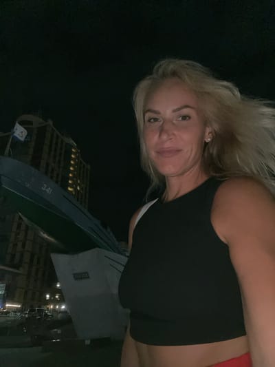 A blonde woman with fair skin and a light complexion is taking a selfie at night. She has light brown eyes and is wearing a black sleeveless top and red bottoms. Her hair is a light blonde and appears windswept. In the background, a building with illuminated windows stands tall against a dark night sky. To the left of the building, a portion of a large, dark green object, possibly a boat or a ship, is visible with the number "341" painted on it. The scene appears to be in an urban or harbor area. The location is not Varnavinskoye, Russia.