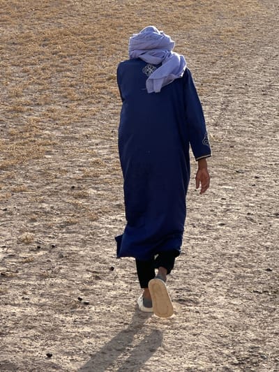 A man wearing a dark blue traditional Moroccan djellaba and light blue cheich is lying on a flat, dry, barren landscape. The ground appears to be a mixture of sand and dry vegetation. The man's head is wrapped in the cheich, and his face is not visible. He is also wearing black pants, gray socks, and white shoes. The location is presumed to be near the city of Sidi Badhaj, Morocco.