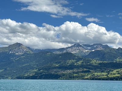 A wide shot captures the serene beauty of Lake Thun in Switzerland, with its clear blue waters gently lapping against a backdrop of majestic mountains. The water, a vibrant turquoise, occupies the foreground, reflecting the bright sky above. White sailboats can be seen dotting the lake, adding a touch of activity to the peaceful scene.

The midground is dominated by lush, green rolling hills that rise to meet the imposing mountains. The slopes are dotted with scattered houses and small villages, their red roofs contrasting with the verdant landscape. Dense forests of dark green trees cover large portions of the hillsides, giving way to grassy meadows in some areas.

The background is a dramatic panorama of rugged mountains, their peaks partially obscured by fluffy white clouds. Patches of snow linger on the higher elevations, hinting at the altitude and the cooler climate. The sky is a brilliant blue, streaked with wispy white clouds that add depth and texture to the celestial canvas. The overall impression is one of tranquility and natural grandeur, a quintessential Swiss alpine vista.