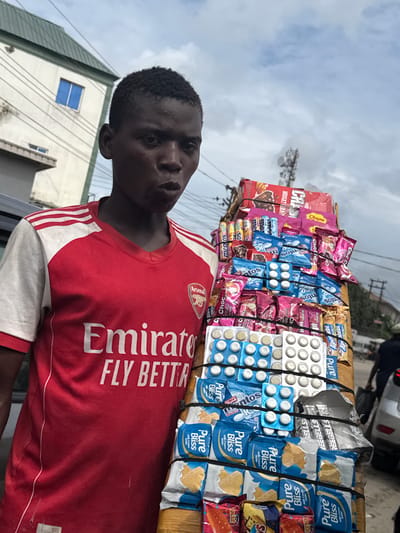 An outdoor, eye-level, full-shot photograph depicts a young Black male standing with a large display of assorted candies and snacks on his left. The male is positioned to the left side of the frame and is looking directly at the camera with a surprised or curious expression. He is wearing a red and white short-sleeved Adidas Arsenal jersey with white stripes on the sleeves and the "Emirates Fly Better" logo printed in white across the chest.
His merchandise is stacked vertically and secured with black elastic bands. The assortment includes various colorful candy wrappers, some in blue and pink, with visible brands like "Pure Bliss" and "Chupa." Small, white blister packs, possibly containing medicine or confectionery, are also part of the display. The background suggests an urban environment in Lagos, Nigeria. To the left, a multi-story building with a green sloped roof and a single blue-tinted window is visible. Power lines crisscross the sky, indicating a developed area. To the right, a silver car and a blurred figure of a person walking away suggest a street setting. The sky is overcast with clouds. The overall impression is that of a street vendor in a bustling urban landscape.