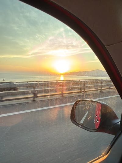 This is a media file showing a view from inside a vehicle, likely a car, looking out the passenger side window. The scene depicts a highway at sunset. The sun is low on the horizon, casting a warm, golden light across the sky and reflecting off the road and the side mirror. The sky is partly cloudy, with wispy clouds catching the sunlight. The road appears to have multiple lanes, and there are some faint streaks on the window, possibly from rain or dirt. In the side mirror, a red car is visible, reflecting the sunset as well. The interior of the car, specifically the roof lining and window frame, is visible in the foreground. The time of day is clearly sunset due to the position and color of the sun. The location is specified as Villeneuve (VD), Switzerland, which suggests the highway is likely near the town. There is no visible text in the media file. The overall mood is serene and peaceful, capturing the beauty of a sunset drive.