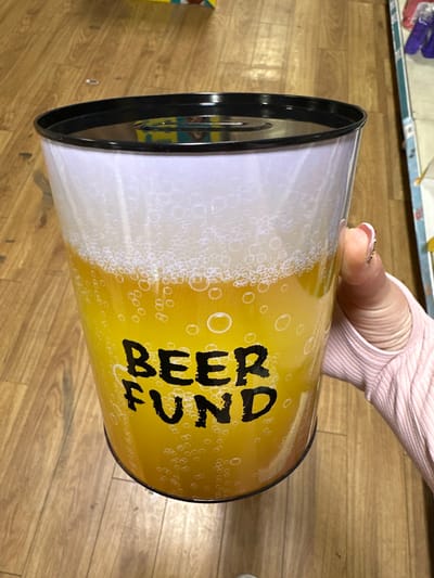 The image shows a close-up of a person holding a novelty beer fund tin can. The tin is designed to look like a pint of beer with foam at the top and bubbles throughout the yellow liquid. The words "BEER FUND" are printed in large, bold, black letters across the front of the can. The person's hand, with fingernails painted, is holding the can. The background is a wooden floor. The image is taken indoors, likely in a domestic or retail setting. There is no identifiable information to suggest the location in the city of Carlow, Ireland.