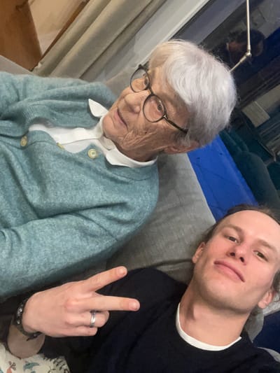 The image shows an elderly woman with short, white hair and glasses, wearing a light blue cardigan over a white collared shirt, seated next to a younger man with long hair who is holding up a peace sign. The two are sitting on a grey couch. The photo appears to be taken indoors. There is a lamp visible in the top left corner of the image. The location of Chambéry, France cannot be determined from this image.