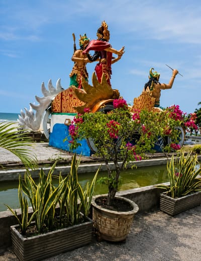 The image depicts a vibrant outdoor scene in Kota Pari, Indonesia. The central focus is a large, ornate sculpture of three figures in traditional attire, appearing to be engaged in a dramatic scene. The sculpture is mounted on a decorative chariot-like structure adorned with golden accents and colorful panels. In the foreground, potted plants, including a tree with pink flowers and several snake plants, are arranged along a concrete walkway. Behind the sculpture, a body of water is visible, reflecting the bright blue sky. The overall environment suggests a public space or a tourist attraction, bathed in sunlight under a clear sky.