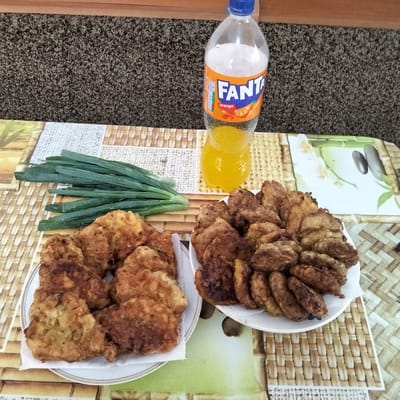 An overhead shot shows a table set for a meal. In the background, a bottle of Fanta orange soda sits next to a carpeted wall. In the foreground, two plates of fried food are arranged on a patterned placemat. To the left, a bunch of green onions lies next to a plate of golden-brown fried food. To the right, another plate is piled high with darker, more irregularly shaped fried food. The setting appears to be indoors, likely a home environment in Cupcini, Moldova, given the available information.