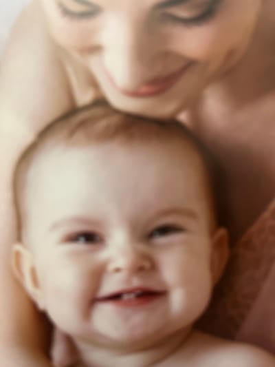 A close-up image, softly blurred, depicts a woman lying down beside a baby. The woman's face, with closed eyes and gently smiling lips, is positioned along the left edge. The baby, partially turned, faces the viewer with bright eyes and a cheerful open mouth revealing tiny teeth. Both figures have light skin tones and are draped in soft fabrics, possibly a blanket or clothing. The warm color palette adds to the tender and intimate nature of the scene.