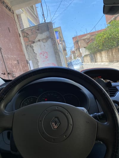 The image is a point-of-view shot from inside a car, looking forward through the windshield. The steering wheel and dashboard are prominent in the foreground, with a Renault logo visible on the steering wheel's center. The car's digital display shows the time as 13:49. Outside the car, a narrow street is visible, lined with buildings that appear to be in a state of disrepair, with exposed brickwork and weathered facades. The sky is blue with scattered clouds, and some power lines are visible. The overall environment suggests an urban setting in a developing area. The presence of graffiti on some of the walls indicates local activity. The specific location is identified as Sidi Moussa, Algeria.