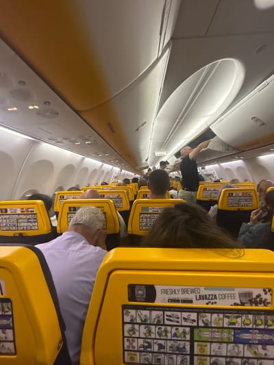 The image depicts the interior of a Ryanair aircraft, likely during a flight. Passengers are seated in rows with yellow headrests, each featuring safety instructions and advertisements for Lavazza coffee. One passenger is standing in the aisle, reaching towards an overhead compartment. The Ryanair logo is visible on the overhead panel. The location of Luqa, Malta, cannot be determined from this image.