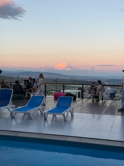A group of people are gathered on a wooden deck overlooking a valley at sunset. The sky is painted with soft hues of pink and orange, with wispy clouds and power lines stretching across the frame. The setting appears to be a rooftop terrace or balcony, as evidenced by the swimming pool on the right side of the image and the lounge chairs arranged on the deck.

Several individuals are seated at tables, engaged in conversation or relaxation. One woman in a white outfit stands and leans towards a table where others are seated. Another group is seated on lounge chairs, some appearing to be looking out at the view. The overall atmosphere suggests a social gathering, perhaps a dinner or an evening get-together, taking place in a scenic location. The time of day is clearly sunset, creating a warm and serene ambiance. The weather appears to be clear, allowing for an unobstructed view of the landscape and the colorful sky. The presence of the pool and lounge chairs suggests a resort or a private residence with recreational amenities. No visible text is present in the image.