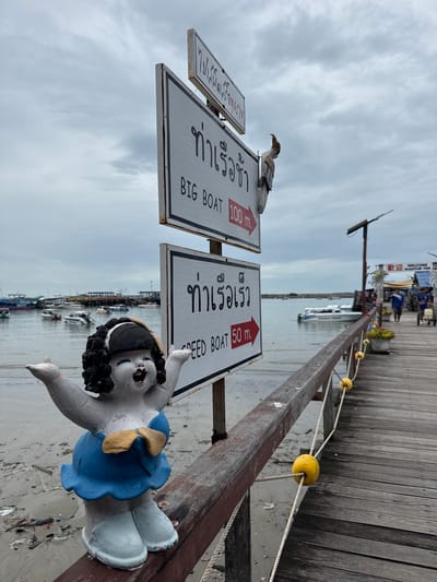 A statue of a joyful young girl in a blue dress stands on a wooden pier in Ban Phe, Thailand, with her arms outstretched. Behind her, a signpost directs visitors to "BIG BOAT" 100 meters to the right and "SPEED BOAT" 50 meters to the right. The pier extends into the water, where various boats are docked. The sky is overcast, and the water is calm. In the background, a sign reads "WELCOME NUANTHIP PIER," indicating the location. The scene is a typical harbor setting, bustling with activity and maritime elements.