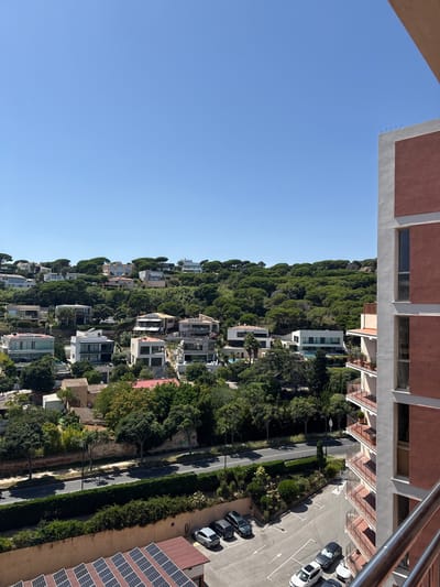The image shows a hillside covered with lush green trees and several modern houses nestled among them. The houses are built with clean lines and large windows, some featuring balconies and swimming pools. A road winds its way up the hillside, with parked cars visible on one side. The scene is bathed in bright sunlight, suggesting it is a clear, sunny day. Solar panels are visible on the roof of a building on the right side of the image. The sky is a clear, vibrant blue. The overall impression is one of a peaceful and affluent residential area in a natural setting. The location context of Sant Feliu De Guíxols, Spain, suggests a coastal Mediterranean environment. There is no visible text in the image.