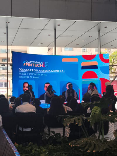 Here is a detailed description of the image:
**General Scene:** The image captures a conference or presentation setting, likely related to fintech, in Guatemala City, Guatemala. The shot is taken from the audience's perspective, looking towards a large screen and a panel of speakers.
**Environment:** The scene appears to be indoors but with access to natural light, as evidenced by the building visible through the large glass panel behind the screen. The setting seems modern, indicated by the design of the lighting and the architectural structure overhead. Potted plants in the foreground add a touch of greenery to the scene.
**People:** A large crowd of attendees fills the seats in the foreground. Many of them are wearing headphones, suggesting simultaneous translation or audio assistance. On stage, there is a panel of speakers, each wearing a headset. At least one of them is speaking into a microphone.
**Objects:** The most prominent object is the large screen displaying the event's title: "Guatemala 20/Fintech 26". The text "Dos caras de la misma moneda: Emisión y aceptación en el ecosistema de pagos" is also visible. There is also a glimpse of a brand logo "Reket" on a part of the screen.
**Actions:** The primary actions occurring are the speakers presenting on stage and the audience listening. The presence of headphones suggests active participation in the event, possibly through translation or interactive elements.
**Interactions:** The main interactions are between the speakers and the audience, facilitated by the microphone and possibly headphones. The screen serves as a visual aid for the presentation.
**Location:** Given the title "Guatemala 20/Fintech 26", it is highly likely that this event is taking place in Guatemala City, Guatemala.