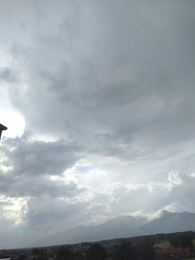 The image captures a panoramic view of a rural landscape under a dramatic, heavily clouded sky. Dense, grey cumulus and stratocumulus clouds dominate the upper two-thirds of the frame, suggesting overcast and potentially stormy weather. Bright patches of sunlight pierce through gaps in the cloud cover, creating visible rays of light that illuminate parts of the sky and landscape below.
In the distance, a majestic mountain range extends across the horizon, with some peaks partially obscured by mist or lower clouds. Below the mountains, a town or village spreads across the mid-ground, characterized by numerous houses with dark roofs and surrounded by abundant green trees and foliage. Patches of open land and fields are also visible. A small section of a dark roof, likely from the structure where the photograph was taken, is present in the top-left corner.
The scene depicts a cloudy day over Gorno Sahrane, Bulgaria, showcasing the interplay of natural light and atmospheric conditions over a mountainous and settled area. No specific human activity or individuals are visible, and there is no discernible text. The time of day appears to be during daylight hours, characterized by diffused light breaking through heavy cloud cover.
