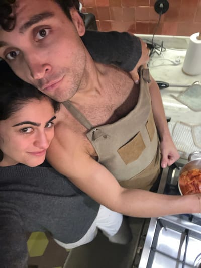 A selfie shows a man and a woman close together. The man, on the left, has dark hair and is wearing a grey sweater on one arm. He has an apron on, revealing a bare chest. The woman is on the right, wearing a grey sweater. In the background, a stovetop with a pot of food is visible, along with kitchen utensils and a tiled floor. The location is Gondarém, Portugal.