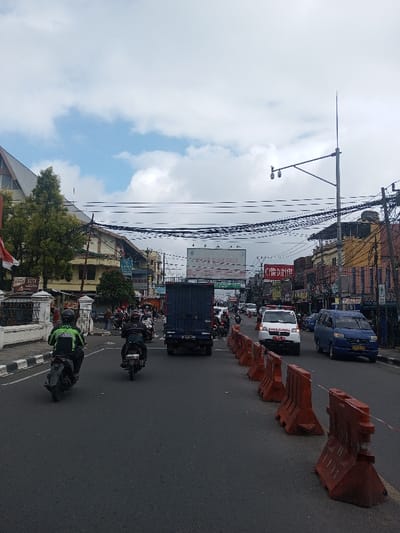 The image displays a daytime urban street scene in Thotagamuwa, Sri Lanka, captured from a ground-level perspective. The sky is partially cloudy with visible patches of blue. The asphalt road is a multi-lane thoroughfare, with a series of orange plastic traffic barriers delineating a section on the right side.
Two individuals on black motorcycles are visible in the foreground, traveling away from the camera. The foremost rider wears a green and black jacket, and the second rider wears a dark jacket. A dark blue box truck follows behind them in the center lane. Further down the road, a white ambulance marked "AMBULANCE" and a blue minivan are observed moving towards the viewer in the rightmost lanes. Beyond these vehicles, moderate traffic consisting primarily of motorcycles and some cars is present.
Multi-story buildings line both sides of the street. On the left, structures include one with a distinctive steep, dark-pitched roof. A white, ornate fence runs along the sidewalk on this side, separating it from the road. Commercial signage, including "COKLAT ELITE," is visible on these buildings. The right side features additional commercial buildings with various advertisements, notably a "CARDINAL" sign. A large billboard displaying "SPACE AVAILABLE" is positioned high above the street in the distance. Numerous black utility lines crisscross the street overhead, supported by utility poles, one of which is equipped with surveillance cameras. Trees and other vegetation are interspersed among the buildings.