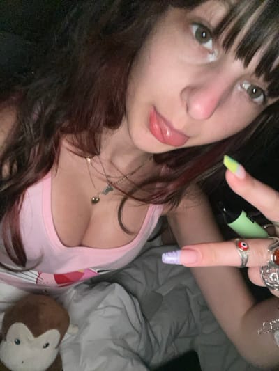 A close-up, high-angle selfie photograph depicts a young adult female, identified as being in Ndlambe Local Municipality, South Africa. She has dark brown hair with bangs and reddish highlights, and light-colored eyes. Her mouth is slightly open with her tongue extended. She wears a light pink scoop-neck tank top, revealing cleavage, with a partially visible graphic that appears to be the Pink Panther. Multiple silver-toned necklaces adorn her neck, one with a heart pendant and another with a small charm. Her right hand is raised, holding a light green cylindrical object resembling a disposable vaping device. Her fingernails are long and manicured; the index finger nail is light purple with a white tip, and a thumb nail has a neon yellow/green tip with a pink line. Several rings are visible on her right hand, including a silver ring with a red "evil eye" symbol on her middle finger, and other ornate silver rings on her ring and pinky fingers. A brown and beige plush monkey toy rests on gray bedding in the lower left foreground. The scene is dimly lit, suggesting an indoor evening setting.