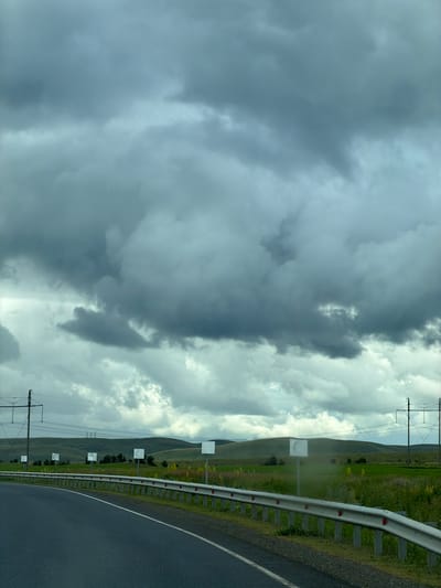 The image shows a road with a metal guardrail, stretching into the distance. The road is paved and appears to be a two-lane highway. To the right of the road is a grassy field with some yellow wildflowers. In the background, rolling green hills are visible. Several white square signs are placed along the side of the road. Power lines and utility poles run across the landscape. The sky is filled with heavy, dark gray clouds, suggesting an overcast or stormy day. There are no people or animals visible in the image. The scene appears to be located in a rural or semi-rural area.