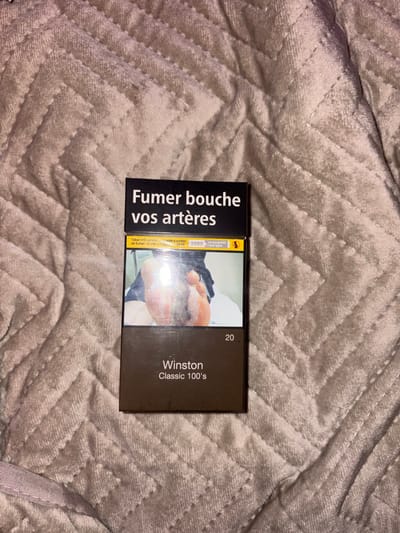 A pack of Winston Classic 100's cigarettes rests on a textured, light-colored blanket. The pack features a dark brown color scheme. The left side of the pack displays the French warning "Fumer bouche vos artères" in bold, black text. A graphic image showing the effects of smoking is visible on the front. The number "20" is printed on the top right, and the brand name "Winston Classic 100's" is printed vertically below it.