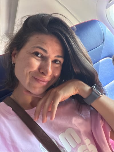 A woman with long, dark hair is taking a selfie in an airplane seat. She is wearing a pink t-shirt and has a brown strap across her chest. She is smiling and resting her chin on her hand. The airplane seat is blue. The image does not provide any information about the city of Donja Cijevna, Montenegro.