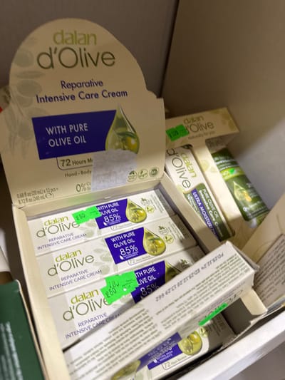 The image shows a close-up, overhead view of several boxes of Dalan d'Olive Reparative Intensive Care Cream, with a price tag of 80 on one of the boxes. The boxes are stacked inside what appears to be a retail display or shipping container. The lighting is artificial, and the focus is sharp on the product packaging. The background is blurred and indistinct, suggesting the image was taken in a commercial setting. There are no people or animals visible in the image. The location is identified as Alakhadzi, Abkhazia.